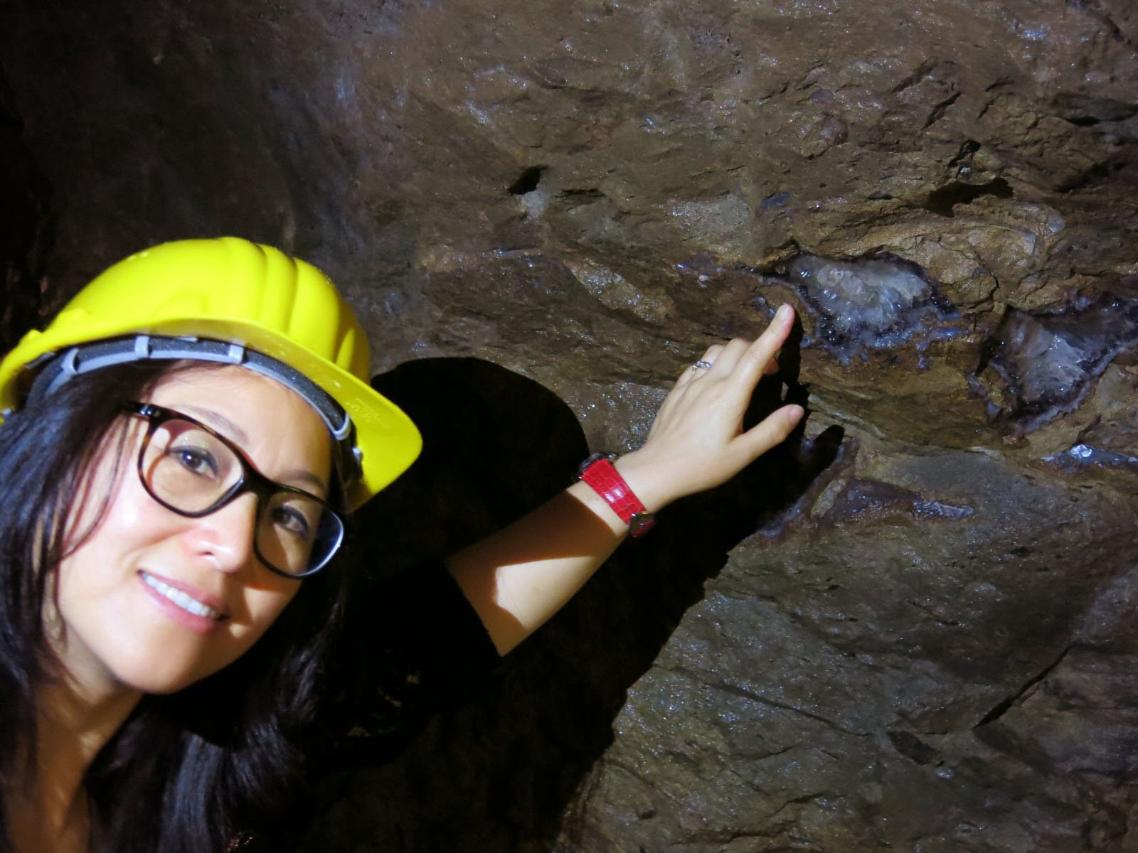 Kehan Li in a yellow hard hat inside a mine on the Yunnan–Myanmar border, hand resting against the rough rock face.